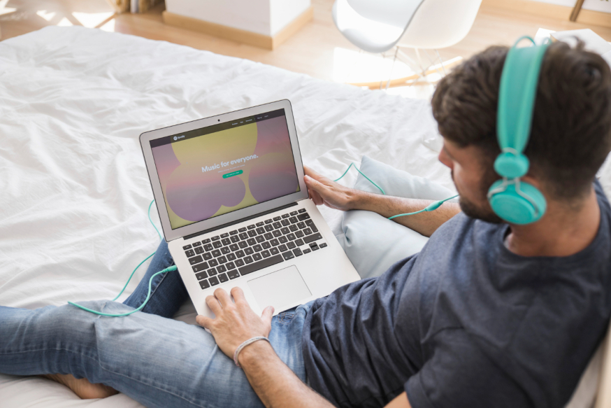 Afbeelding: Een man zit op een bed met een laptop op schoot en een lichtblauwe koptelefoon op, kijkend naar het 'Music for everyone' startscherm van Spotify