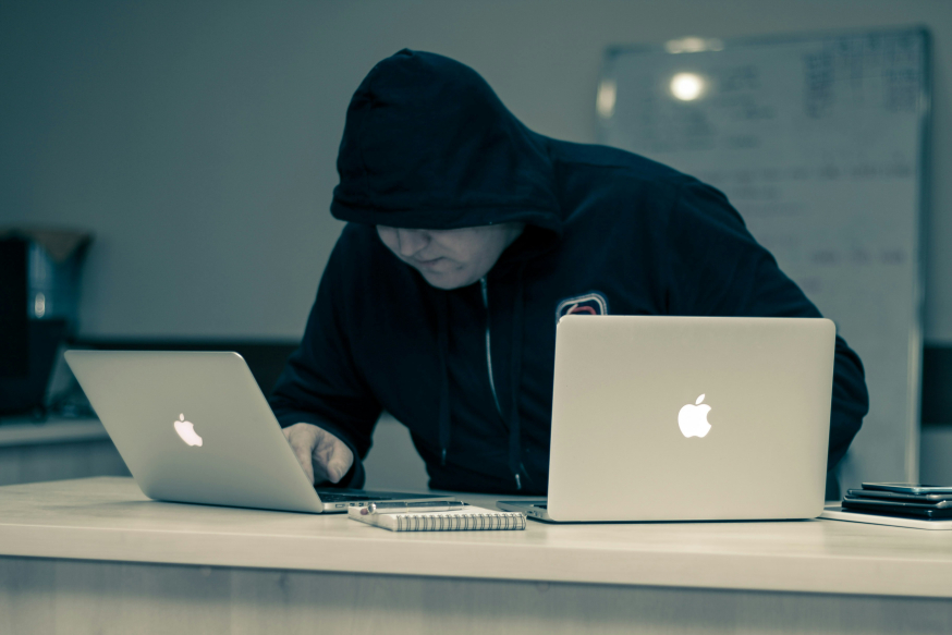 Persoon met capuchon achter laptops, symboliseert anonieme cybercrimineel