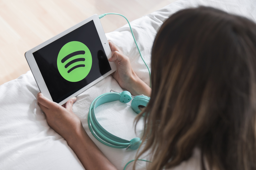 Afbeelding: Een jonge vrouw ligt op een bed en kijkt aandachtig naar een witte tablet met het Spotify-logo, met een lichtblauwe koptelefoon naast haar